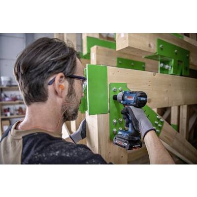 Ein Handwerker befestigt Holzträger mit einem Akku-Bohrschrauber. Er trägt Gehörschutz und Arbeitshandschuhe, arbeitet an einer Holzkonstruktion.