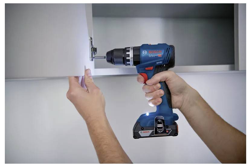 Eine Person verwendet eine blaue und schwarze kabellose Bohrmaschine, um eine Türangel an einer weißen Schranktür zu befestigen und veranschaulicht damit Heimwerkerarbeiten.