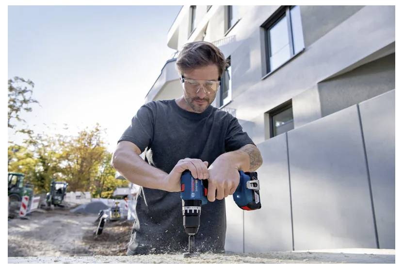Eine Person mit Sicherheitsbrille verwendet einen blau-roten Akkuschrauber auf einer Baustelle mit Gebäuden und Bäumen im Hintergrund.