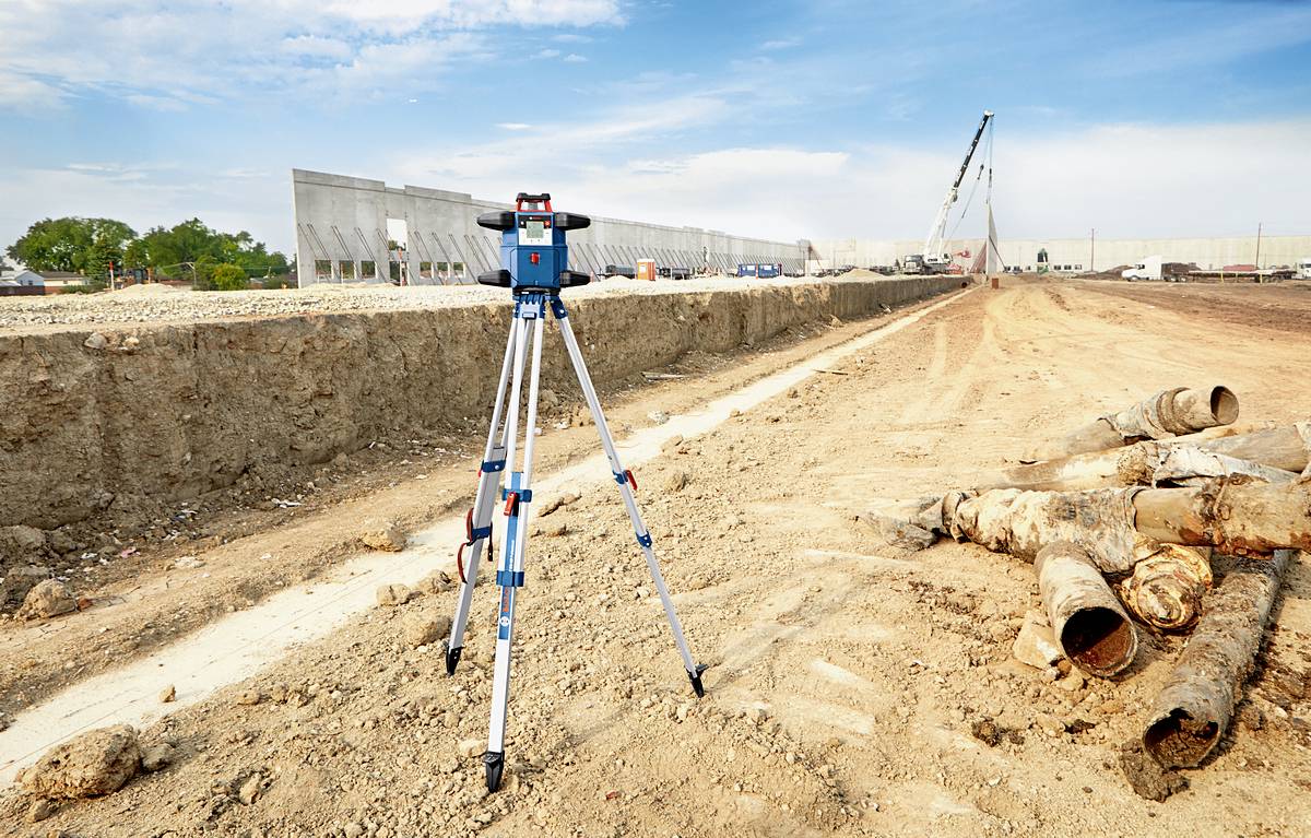 'Baustelle mit Vermessungsgerät im Vordergrund. Ein großer Bagger und Rohrleitungen in der Nähe. Klare, sonnige Wetterbedingungen.'
