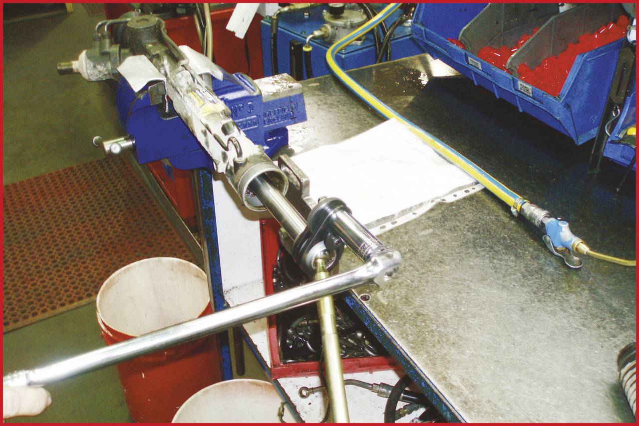 Ein Mechaniker zieht eine Schraube an einem Metallgerät in einer Werkstatt fest. Im Hintergrund sind Werkzeuge und Schläuche sichtbar.