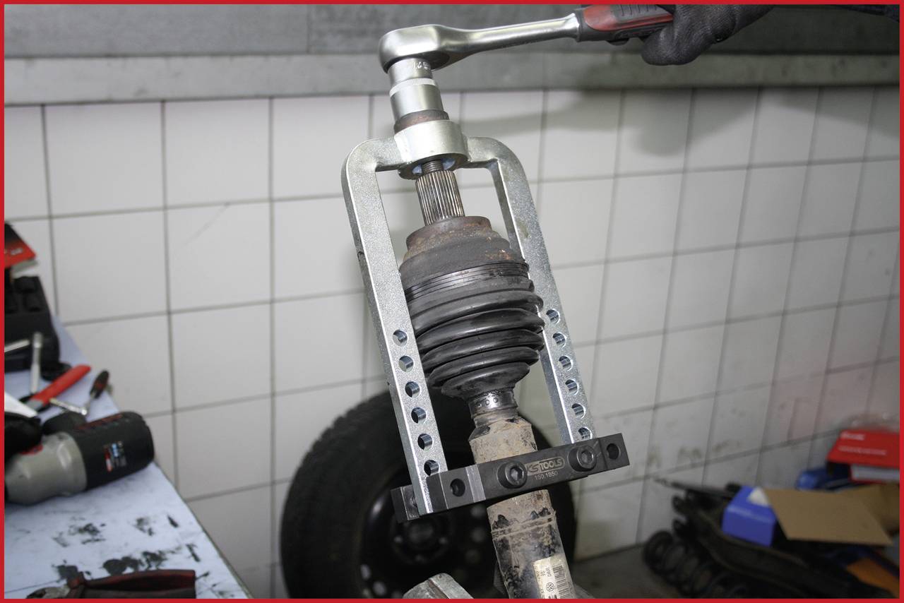 Ein Mechaniker entfernt ein Antriebswellengelenk mit einem Abzieherwerkzeug. Arbeitsplatz in einer Autowerkstatt sichtbar im Hintergrund.