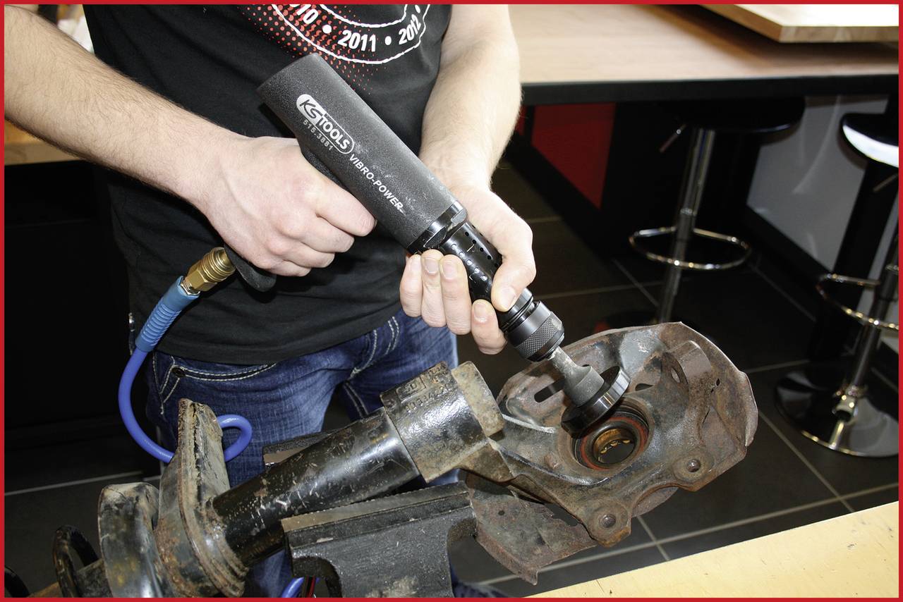 Eine Person bearbeitet ein Autoteil mit einem Druckluftwerkzeug. Werkzeuge und Autoteil sind auf einer industriellen Werkbank platziert.