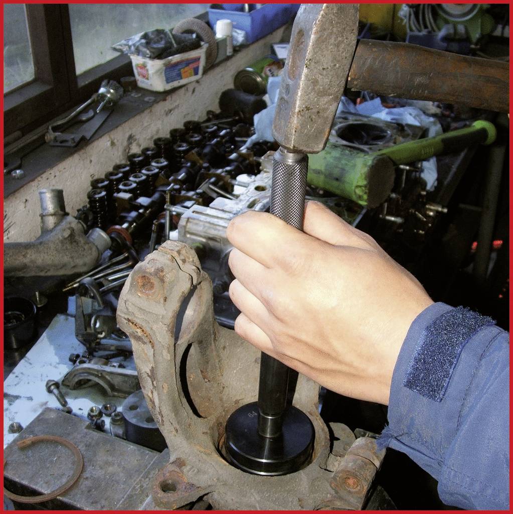 Eine Person benutzt einen Hammer und ein Werkzeug, um ein Autoteil in einer Werkstatt zu montieren. Im Hintergrund liegen Autoteile.