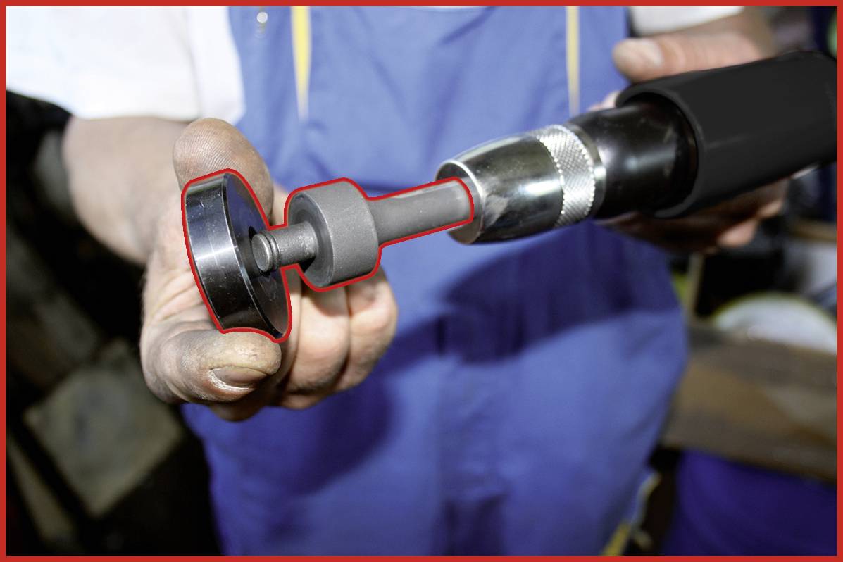 Eine Person hält ein rot markiertes Werkzeug, das wie ein Poliergerät aussieht. Hintergrund zeigt eine Werkstattumgebung.