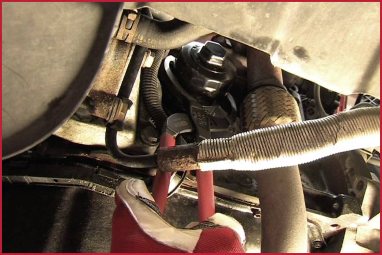 Unterseite eines Fahrzeugs mit Fokus auf eine Hand in einem roten Handschuh, die unter der Motorhaube an Kabeln arbeitet.