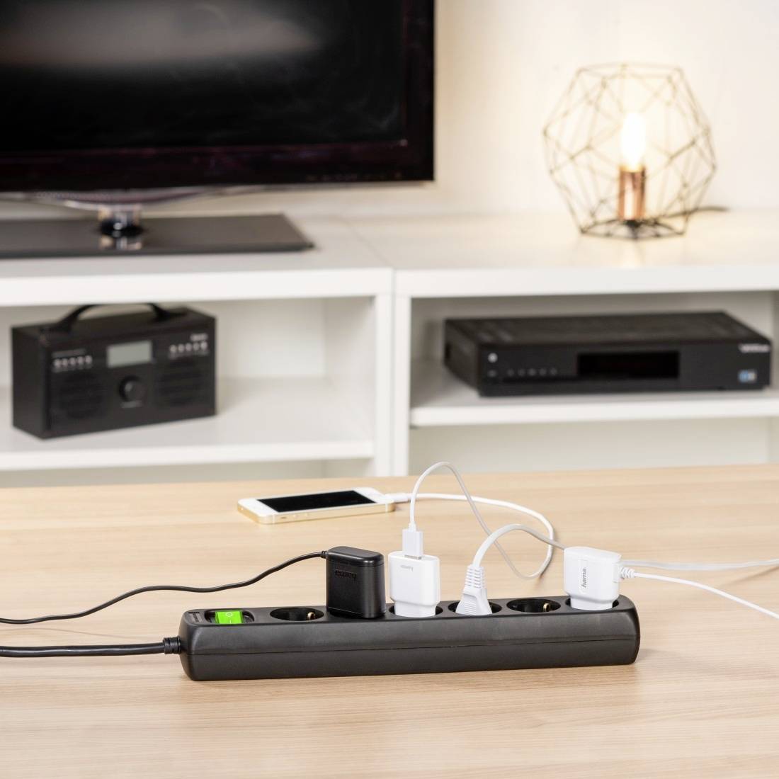 Ein Tisch mit einer Mehrfachsteckdose, an der vier Geräte angeschlossen sind, darunter ein Smartphone-Ladegerät und ein Lampenkabel. Im Hintergrund stehen ein Fernseher und ein Radio.