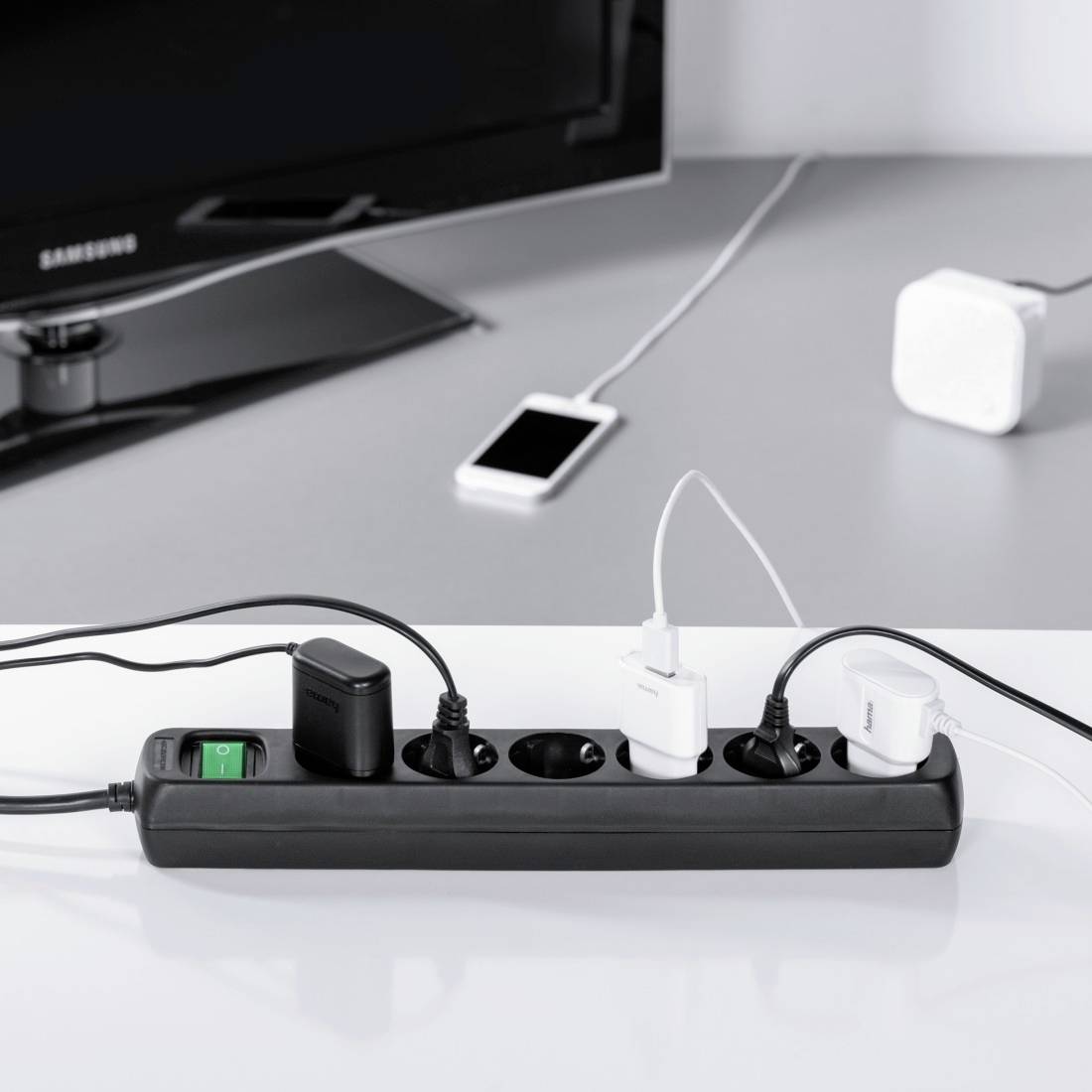 Steckdosenleiste mit mehreren angeschlossenen Ladegeräten; im Hintergrund ein Smartphone, Fernseher und Router.