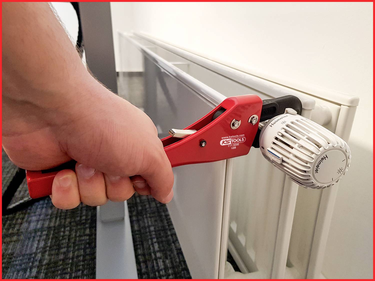 Eine Hand benutzt eine rote Zange, um das Thermostatventil eines Heizkörpers zu drehen. Ein moderner Büroraum ist im Hintergrund sichtbar.