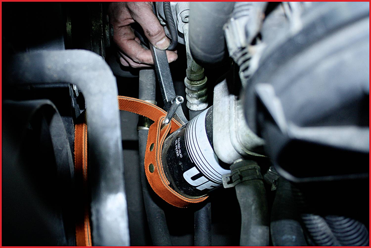 Eine Hand benutzt ein Werkzeug, um einen Ölfilter in einem Motorraum zu lösen. Der orangefarbene Riemen spannt sich um den Filter.