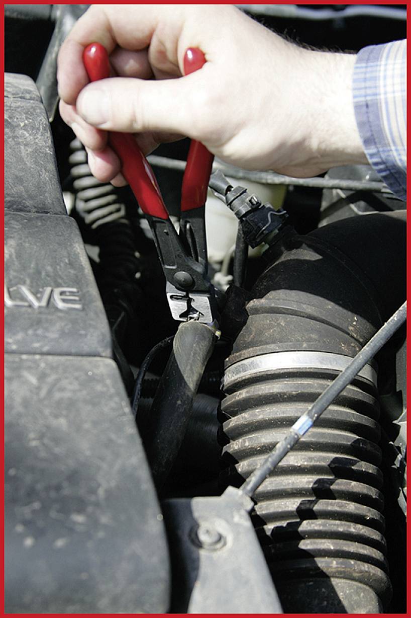 Eine Hand hält eine rote Zange und schneidet ein Kabel im Motorraum eines Autos ab.