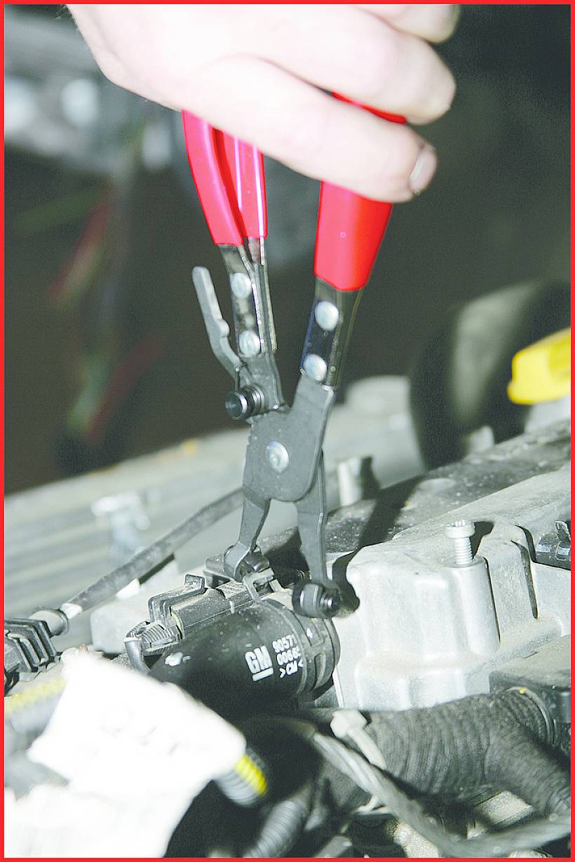 Eine Hand hält eine rote Zange, die ein schwarzes Bauteil in einem Motorraum bearbeitet.