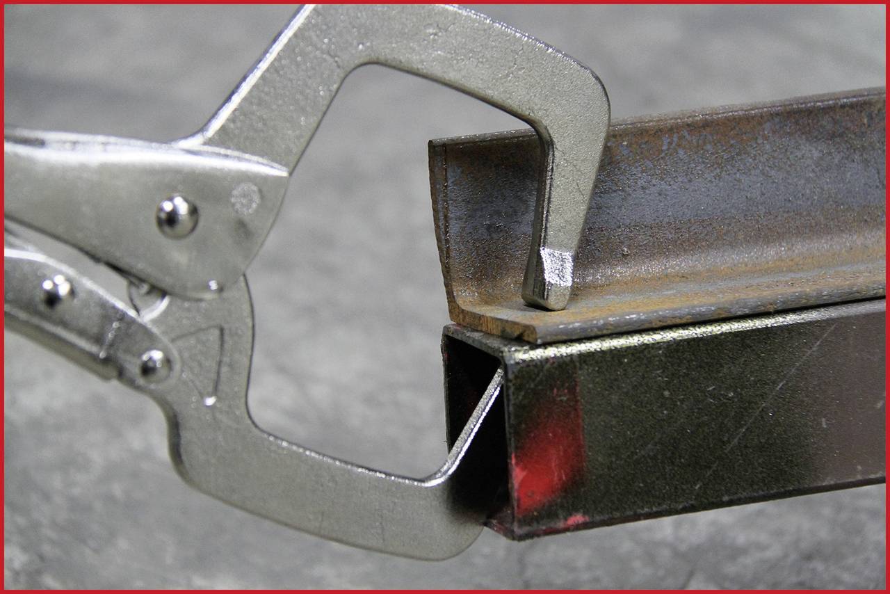 'Ein Metallgreifer hält ein L-Profil gegen ein Vierkantrohr. Beide Gegenstände aus Metall. Industrialle Umgebung.'