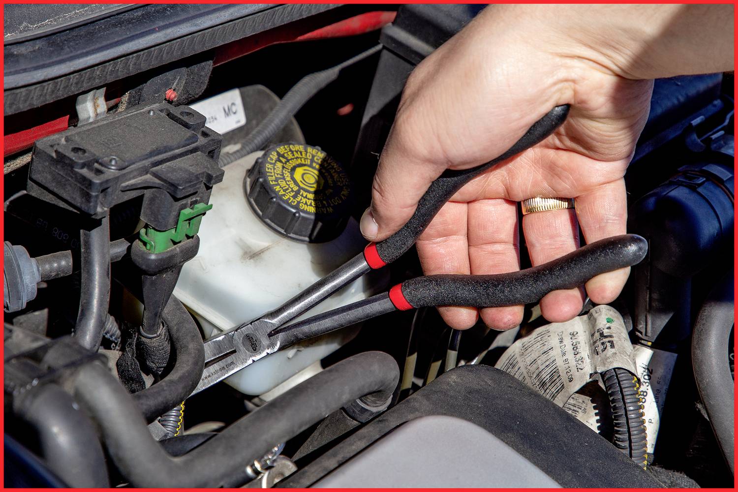 Eine Hand hält eine Zange und arbeitet an einem Automotor. Ein Kühlflüssigkeitsbehälter ist sichtbar.