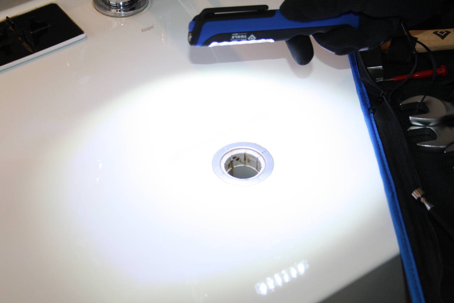Eine Person leuchtet mit einer Taschenlampe in ein Waschbecken, in dem der Abfluss sichtbar ist. Auf der dunkel gehaltenen Seite liegt zusätzliches Werkzeug.