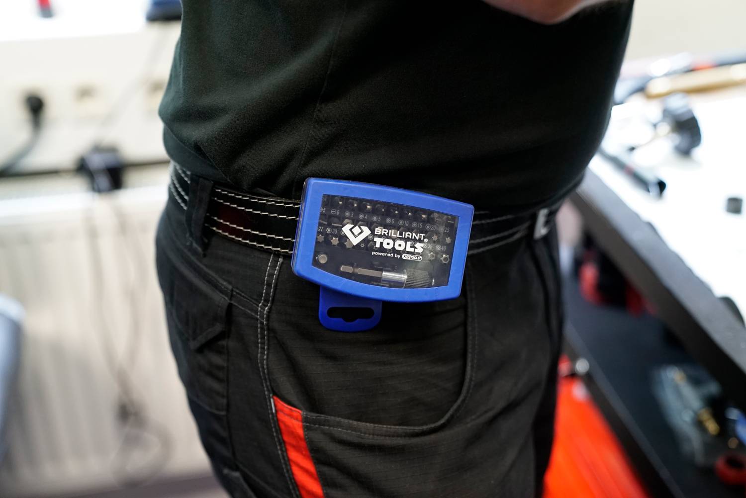 Eine Person trägt ein blaues 'Brilliant Tools'-Multimeter am Gürtel. Im Hintergrund sind Werkzeuge und Werkstattoberfläche zu sehen.