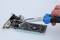 Eine Hand mit blauen Zangen manipuliert eine Computer-Grafikkarte auf weißem Hintergrund.