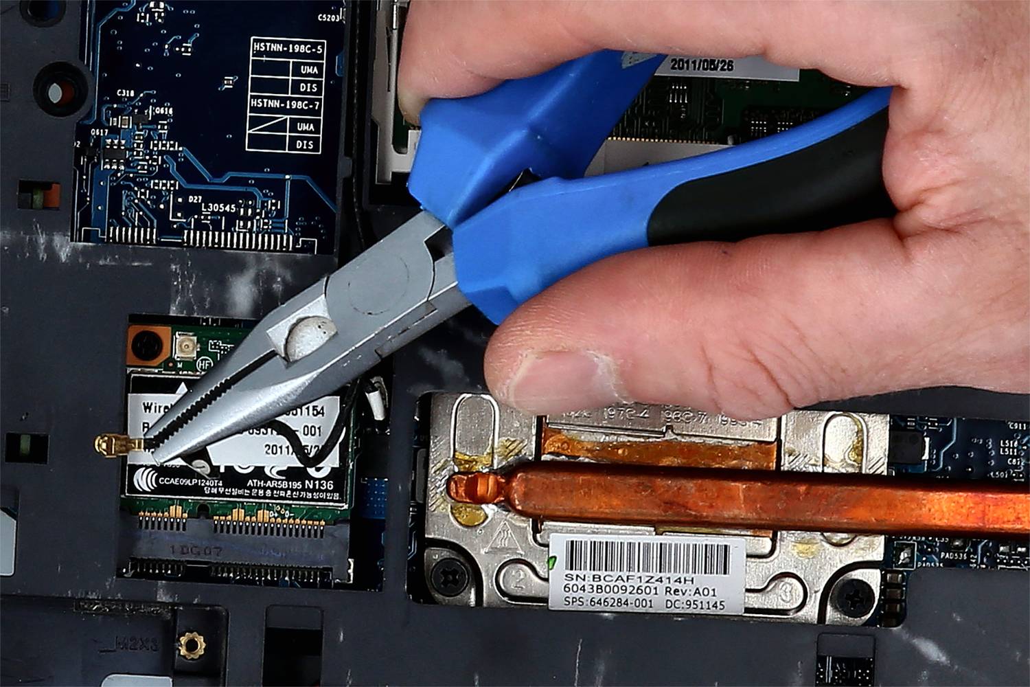 Eine Hand benutzt eine Zange, um einen kleinen goldenen Draht in einem geöffneten Laptop zu manipulieren, nahe einer WLAN-Karte und einem Kühlkörper.