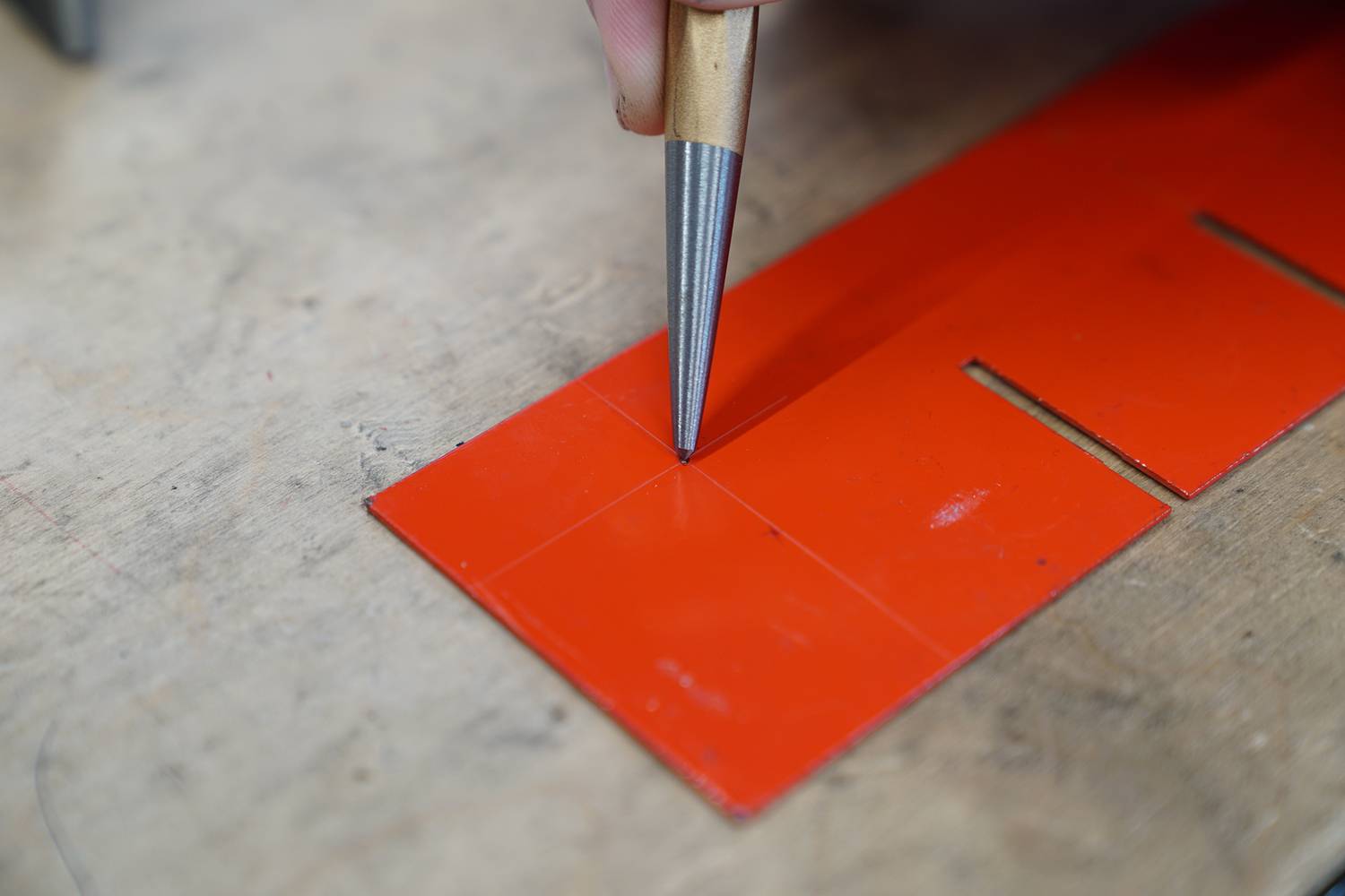 Eine Person markiert mit einem spitzen Werkzeug präzise einen Punkt auf einem roten Plastikbogen, offenbar beim Basteln oder Handwerken.