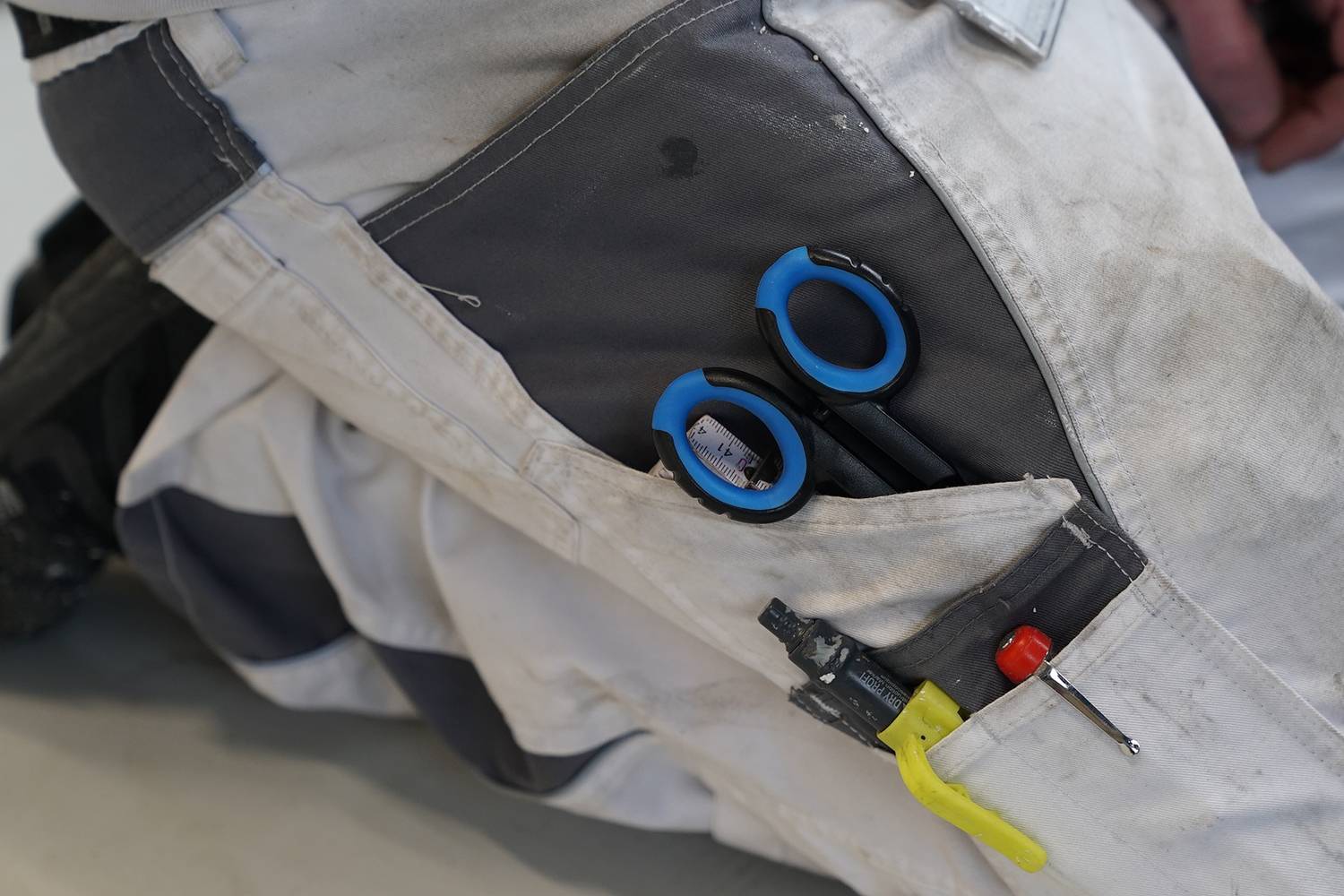 Eine ausgeblichene Arbeitshose mit mehreren Werkzeugen, darunter eine Schere und ein Schraubendreher, in der Vordertasche.