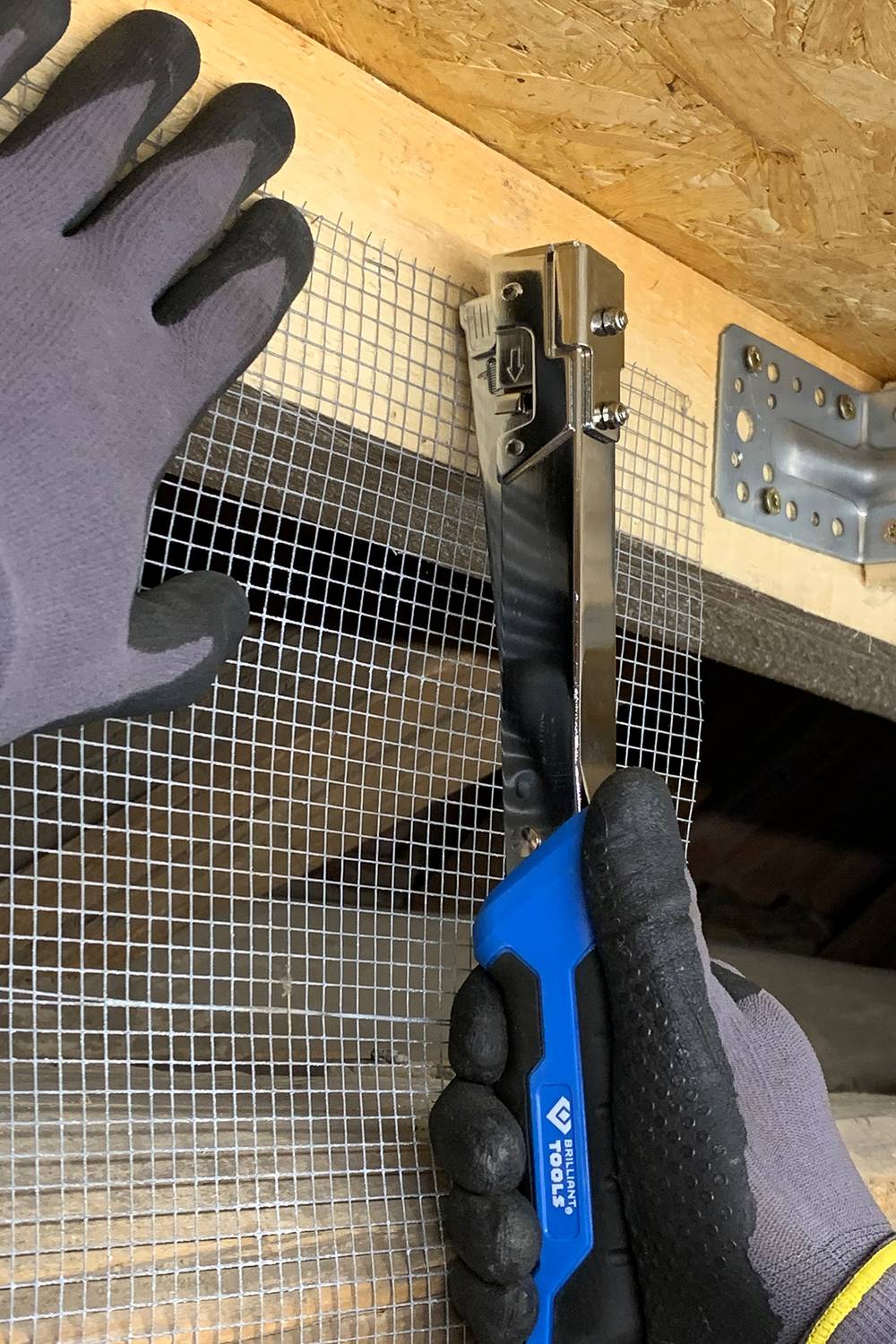 Eine Person befestigt ein Metallgitter mit einem Handtacker an einer Holzoberfläche. Beide Hände tragen Arbeitshandschuhe.