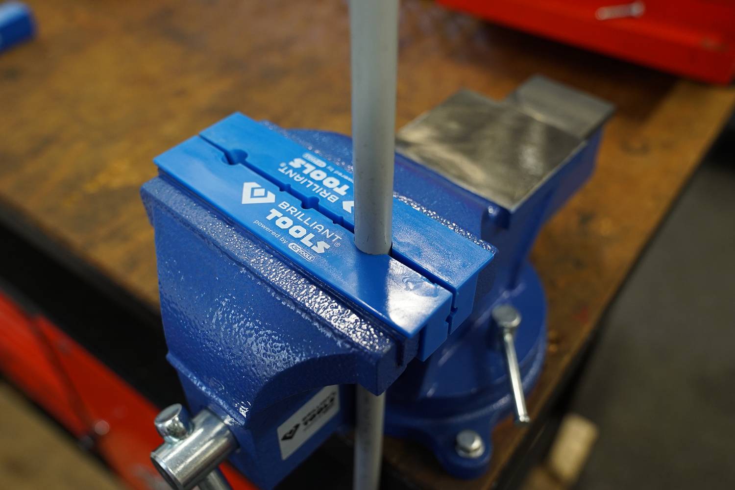 Ein blauer Schraubstock ist fest an einer Werkbank montiert und hält ein weißes Metallrohr. Der Schraubstock zeigt das Logo 'Brilliant Tools'.