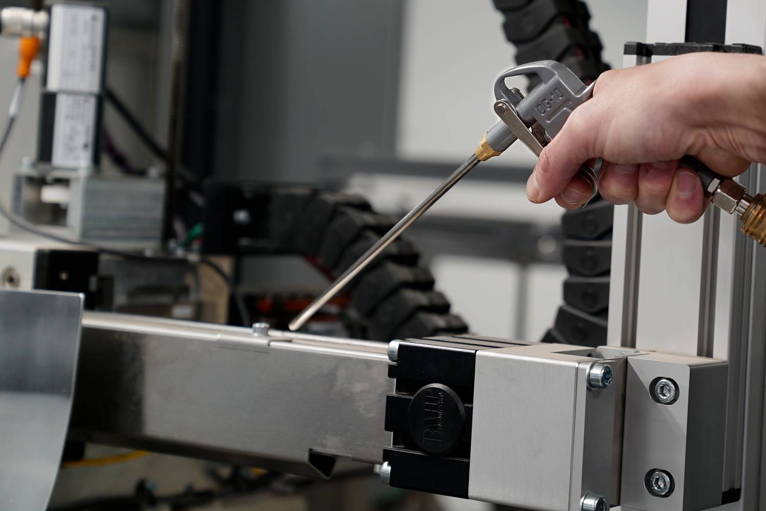 Eine Hand hält eine Druckluftpistole und reinigt eine Maschine. Details der Maschinenbauteile sind sichtbar.