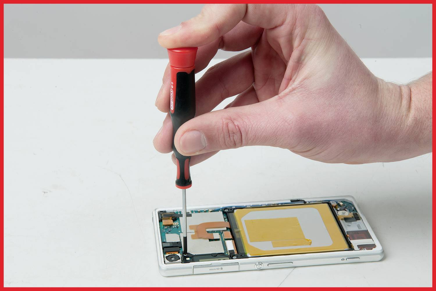Eine Hand benutzt einen Schraubenzieher, um ein Smartphone zu reparieren. Das Telefon liegt offen, und innere Komponenten sind sichtbar.