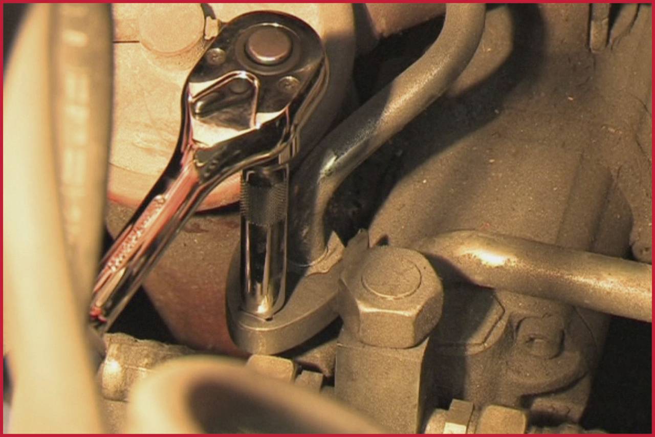 'Ein Ratschenwerkzeug wird verwendet, um eine Schraube in einem Motorraum zu lösen. Der Bereich ist von Rohren und Leitungen umgeben.'