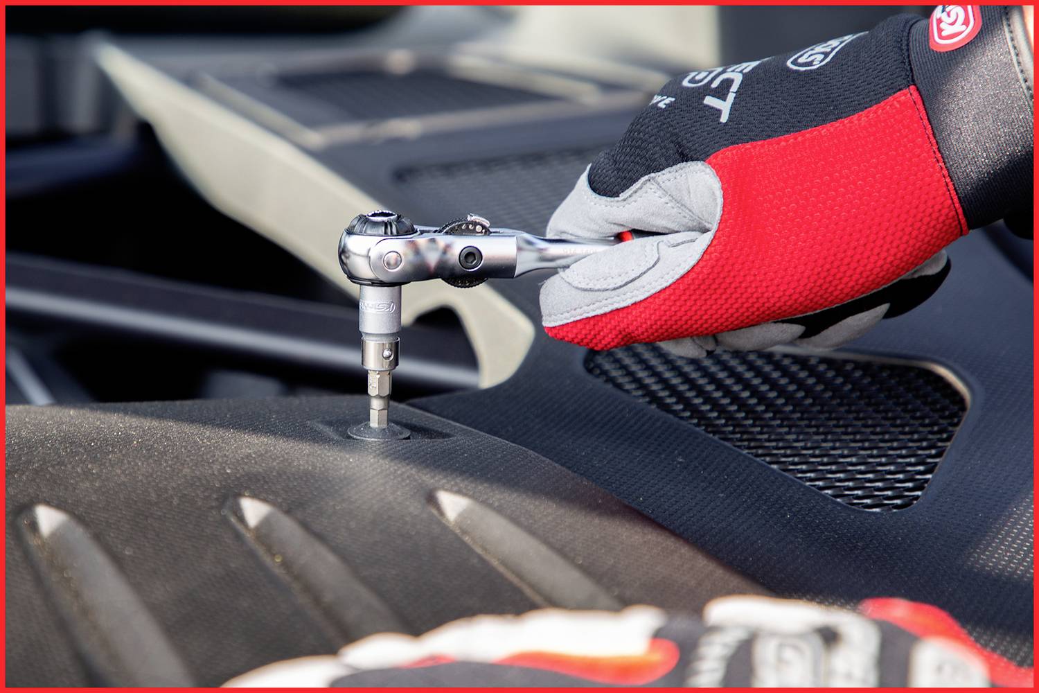 Eine Person mit rot-schwarzer Arbeitshandschuh benutzt eine Ratsche, um eine Schraube unter der Motorhaube eines Fahrzeugs festzuziehen.