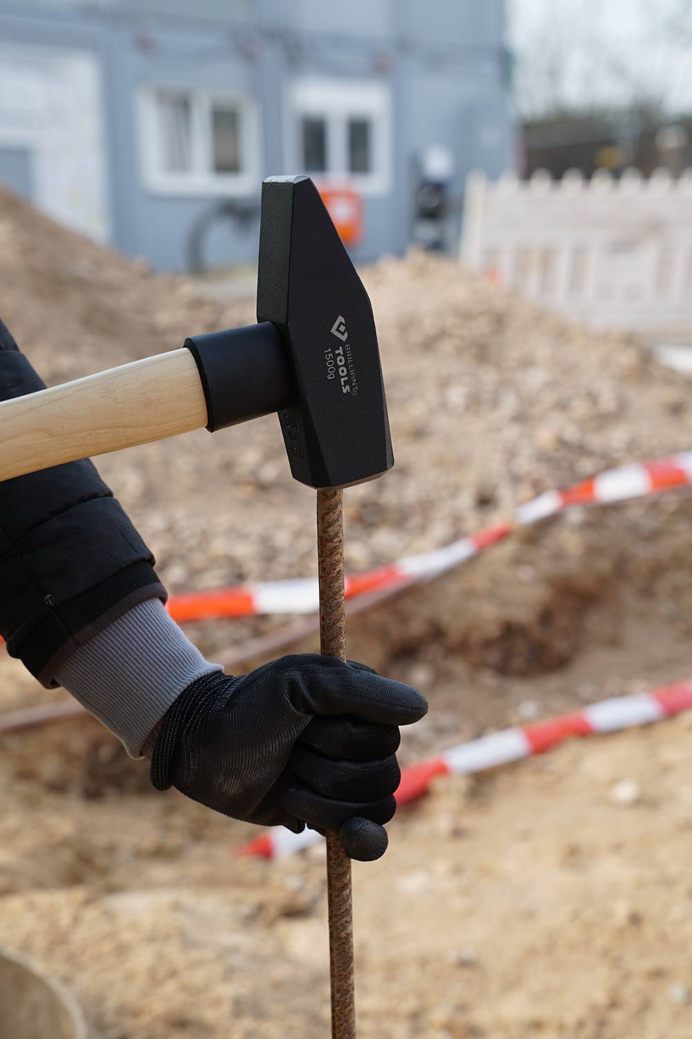 Eine Person auf einer Baustelle schlägt mit einem schwarzen Hammer auf eine Metallstange. Hintergrund zeigt Erdhaufen und Absperrband.