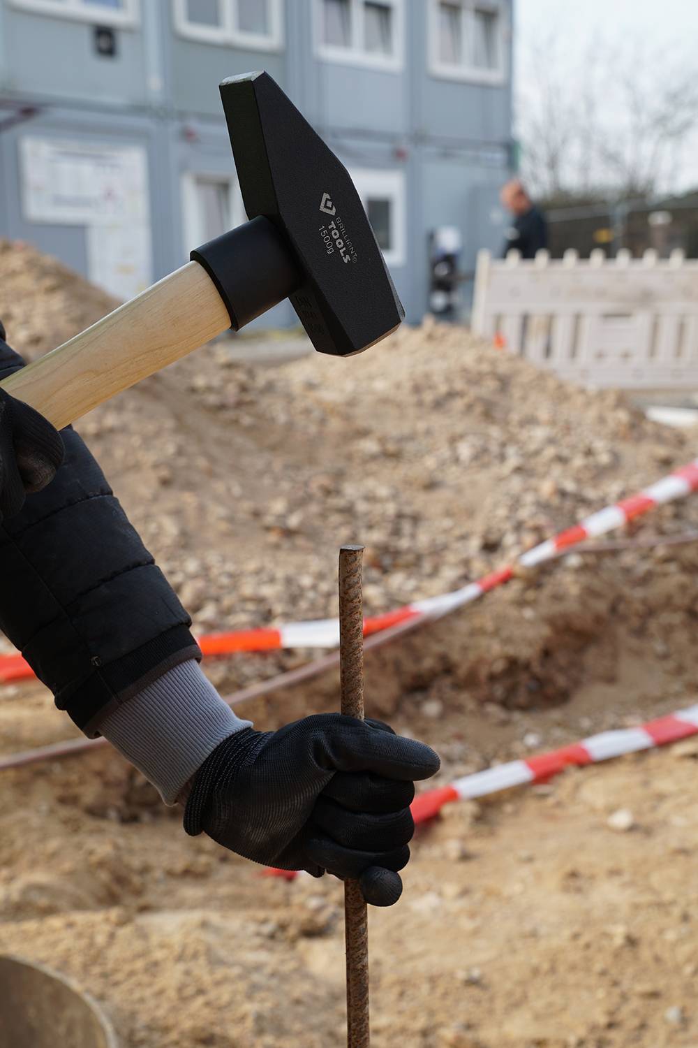 Person hält einen Hammer und schlägt einen Metallstab in den Boden auf einer Baustelle ein, umgeben von Absperrband.