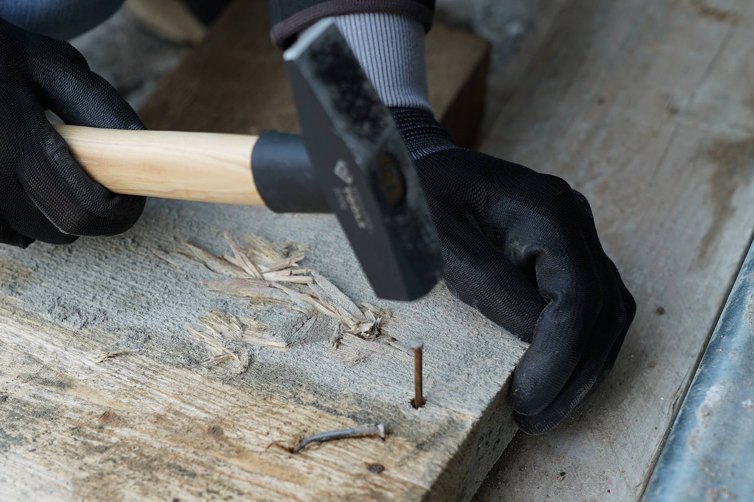 Eine Person schlägt mit einem Hammer einen Nagel in ein Holzbrett. Nägel und Holzsplitter sind sichtbar. Die Person trägt Handschuhe.