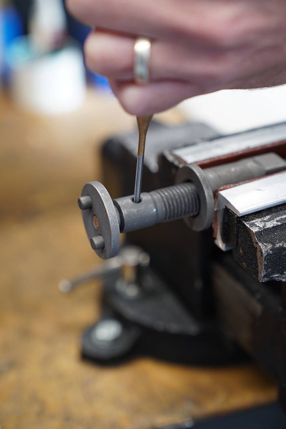 Eine Hand justiert eine Schraube mit einem kleinen Werkzeug in einer Werkstatt. Schraubstock hält das Metallteil fest.