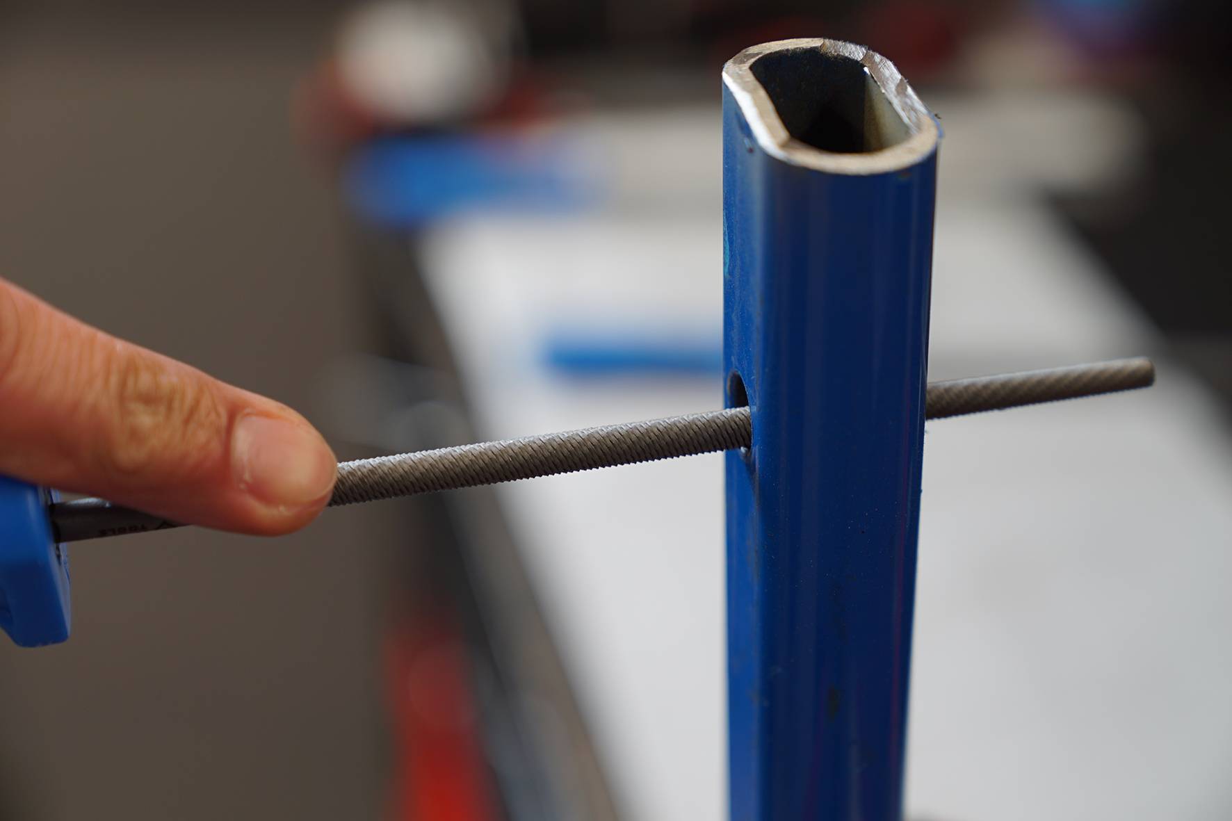 Eine Hand feilt eine runde Öffnung in einem blauen Metallrohr. Im Hintergrund sind unscharfe Werkzeuge zu sehen.