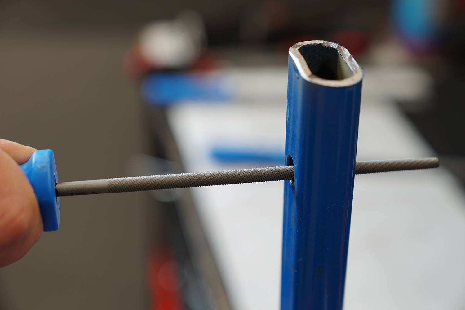 Eine Hand hält eine Feile, die durch ein Loch in einem blauen Metallrohr geführt wird. Die Feile formt oder glättet das Innere des Rohrs.