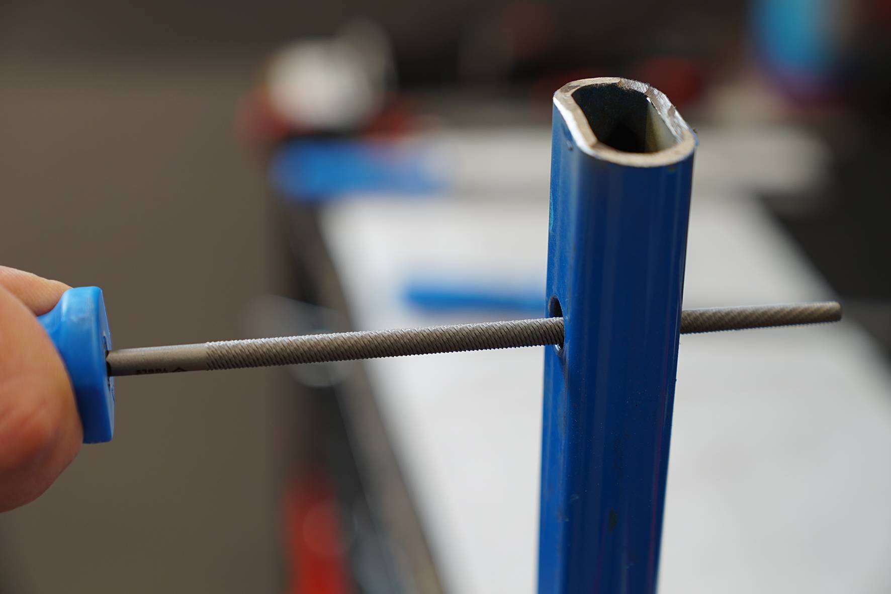 Eine Feile wird durch ein blaues Metallrohr gezogen, um die Kanten im Inneren zu glätten. Einige unscharfe Werkzeuge sind im Hintergrund.