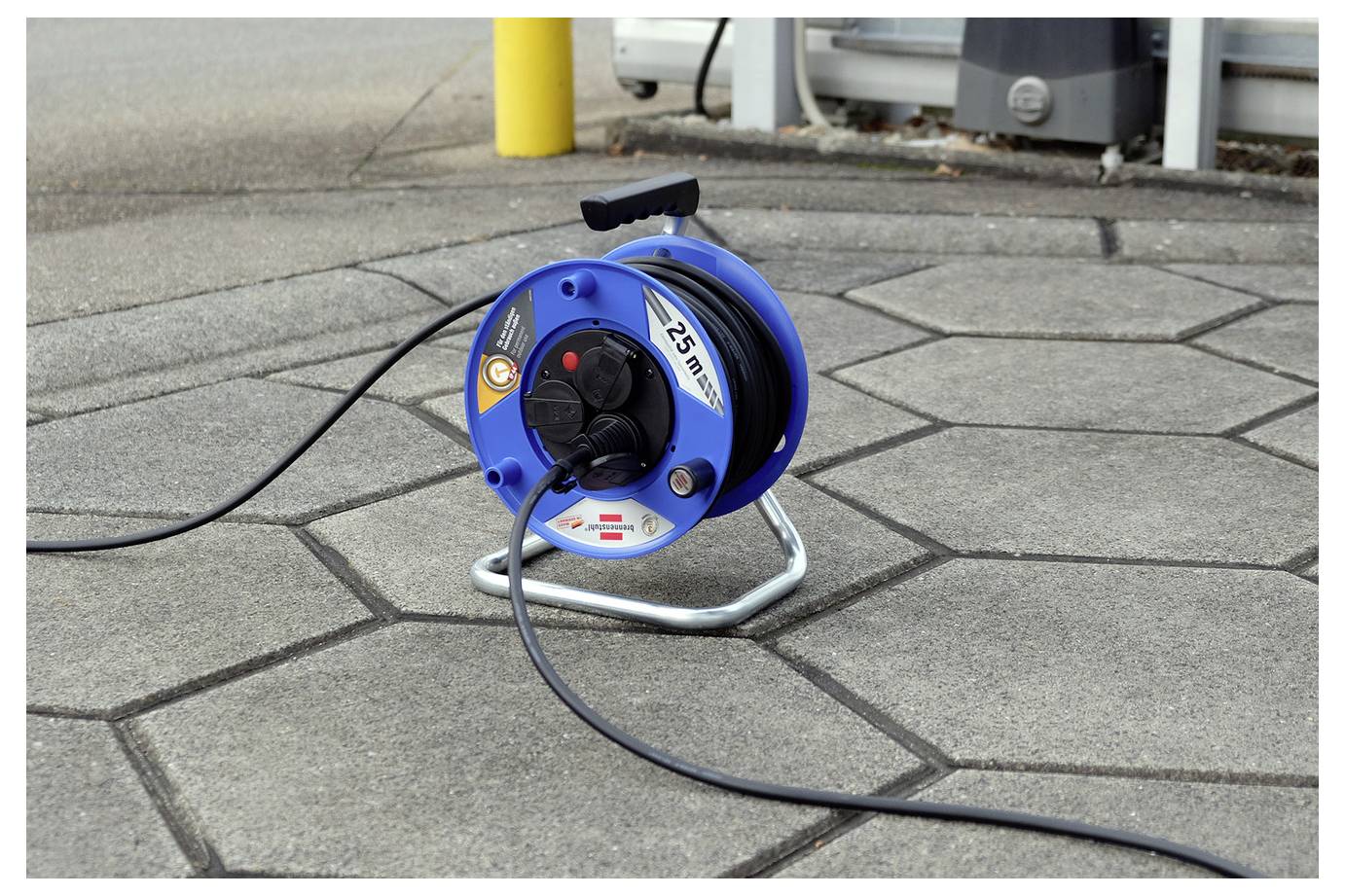 Blaue Kabelrolle mit schwarzem Stromkabel auf einer gepflasterten Fläche im Freien, in der Nähe eines Gebäudes.