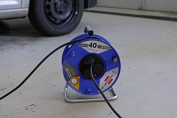 Ein elektrisches Verlängerungskabel, auf einer blauen Kabeltrommel mit 40-m-Markierung, steht auf dem Boden neben einem Autoreifen.