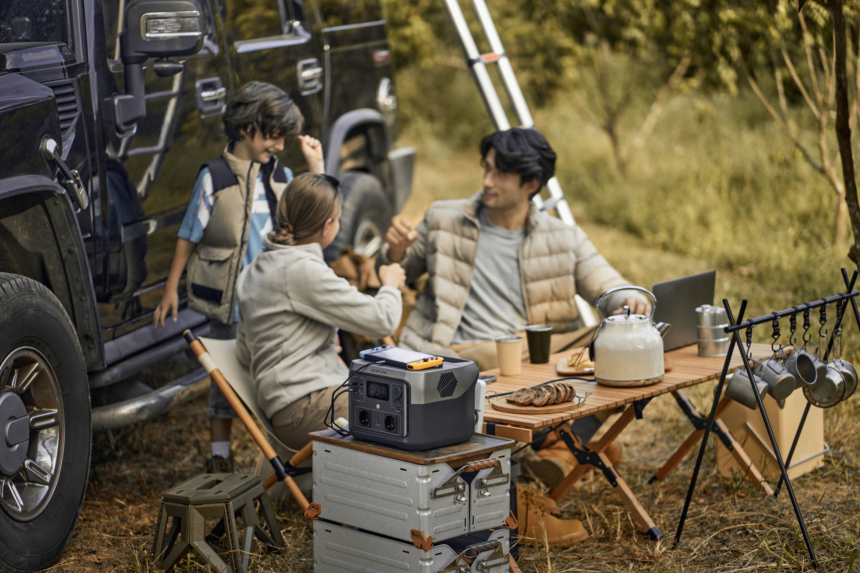Familie sitzt beim Camping an einem Tisch neben einem Auto im Wald. Sie genießen eine Mahlzeit und ein tragbarer Generator steht auf einem Koffer.