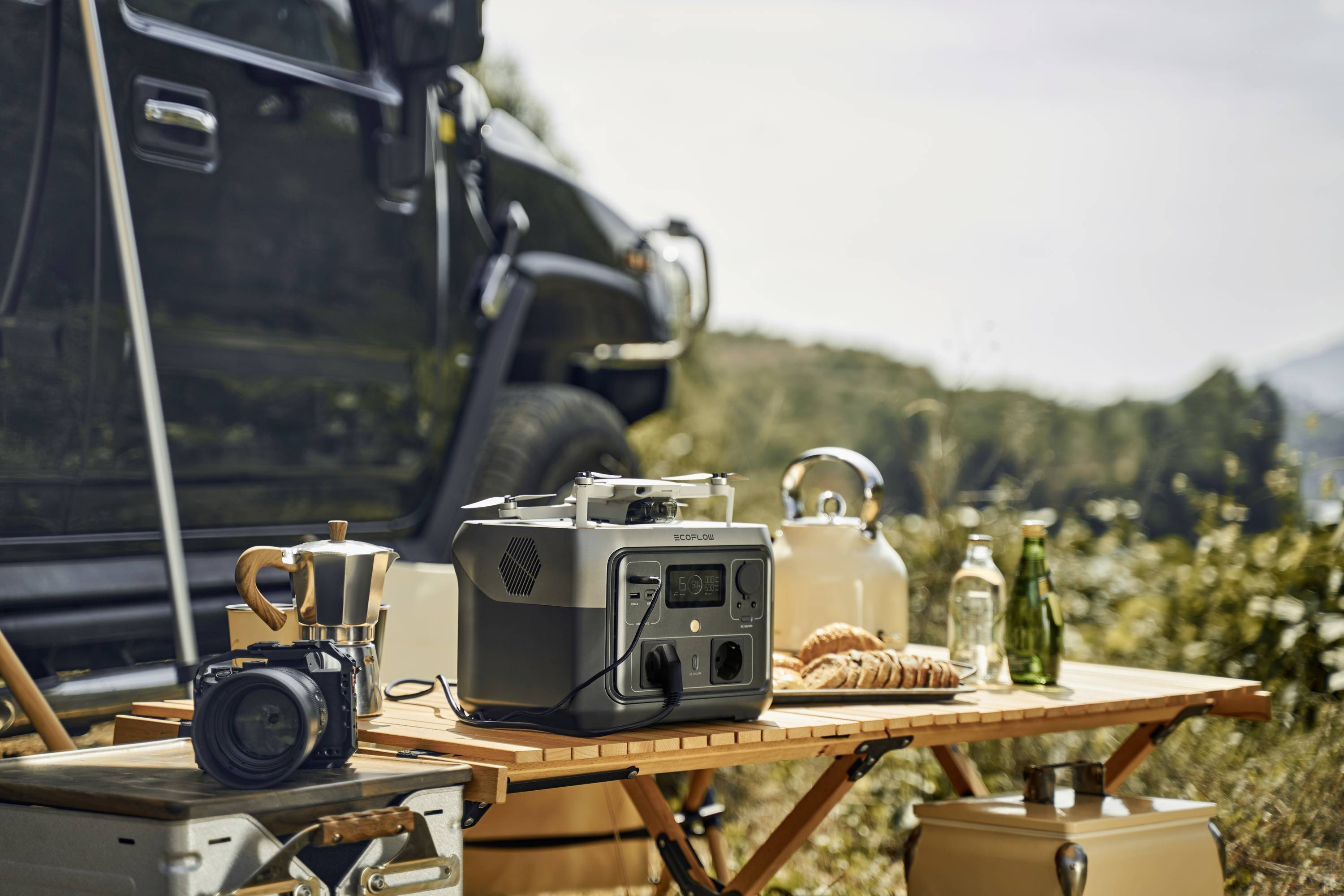 Ein Camping-Tisch neben einem Auto mit einem tragbaren Generator, einer Kamera, Gebäck und Getränken. Hintergrund: Bäume und Berge.