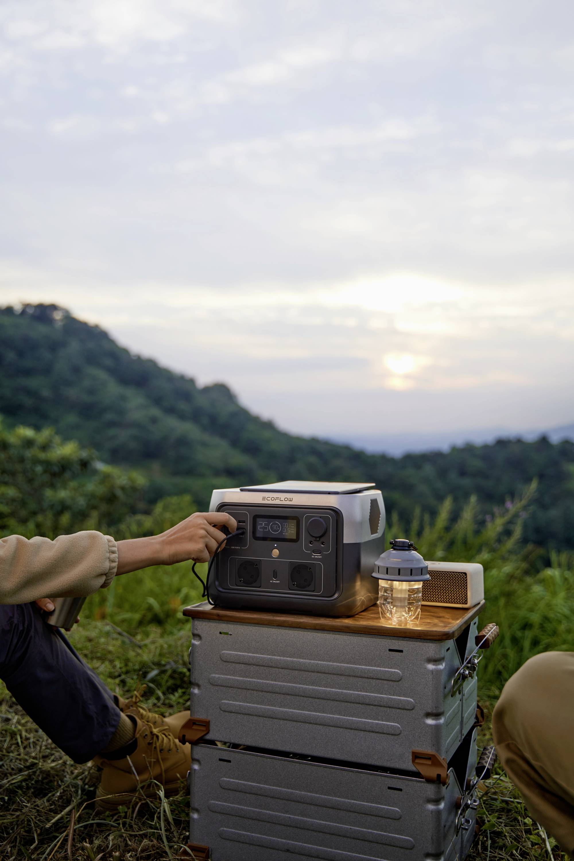 Person auf einem Berg beim Sonnenuntergang, die ein tragbares Solarmodul zur Stromversorgung nutzt, neben einer Laterne auf einer Metallbox.