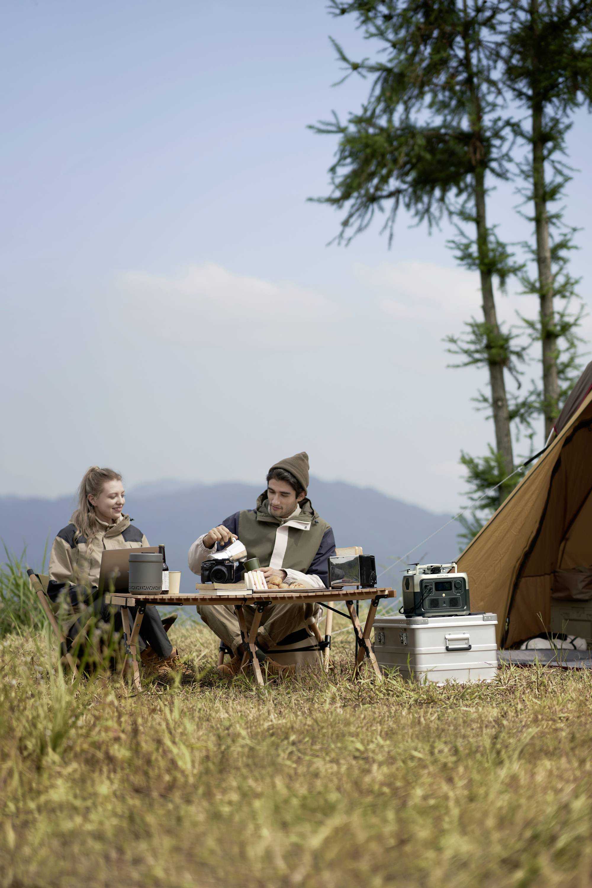 Zwei Personen sitzen beim Camping an einem Tisch im Freien. Eine Person gießt ein Getränk ein. Im Hintergrund ist ein Zelt und Landschaft.