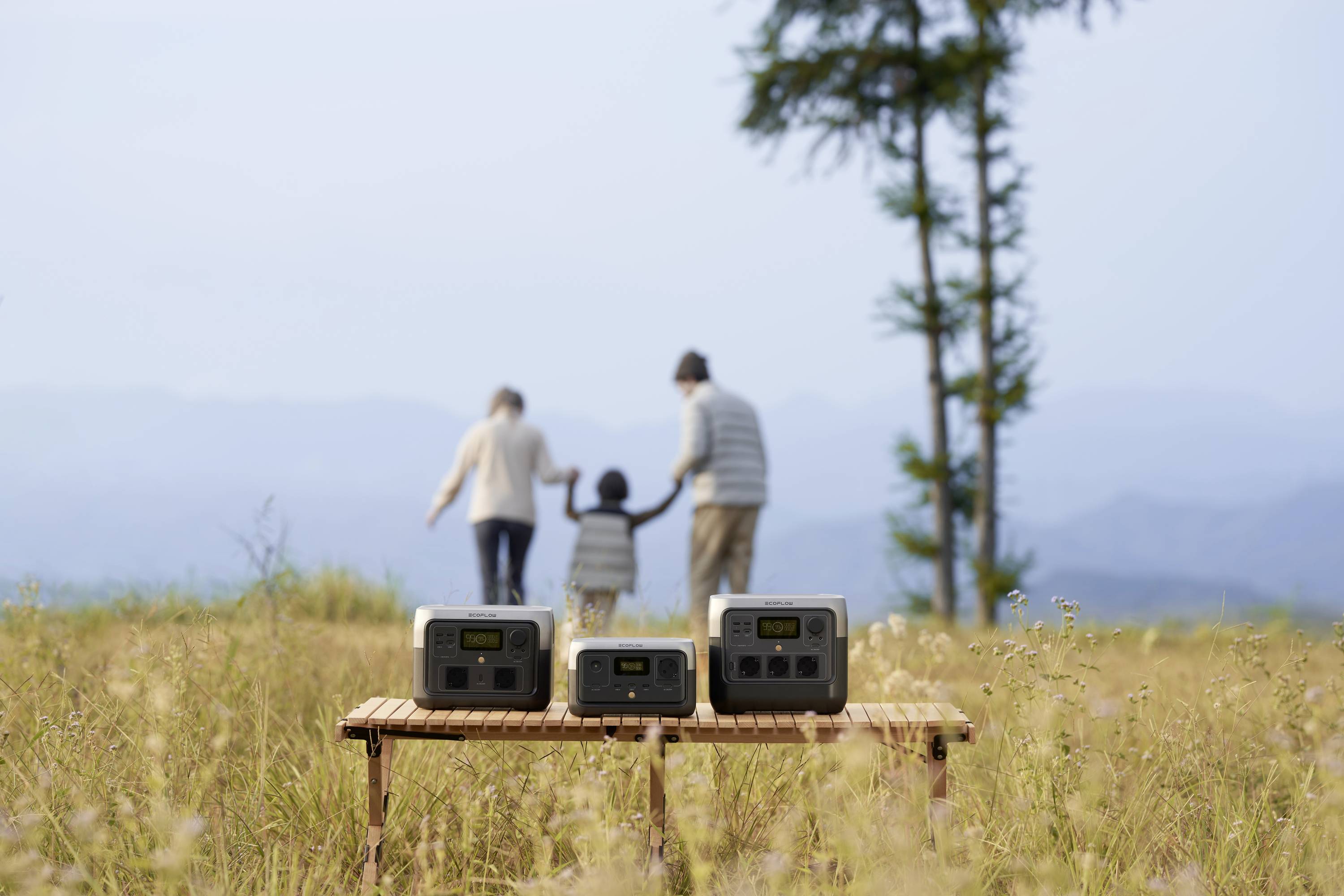 Drei tragbare Stromgeneratoren stehen auf einem Tisch auf einem Feld. Im Hintergrund hält eine Familie an der frischen Luft Händchen.