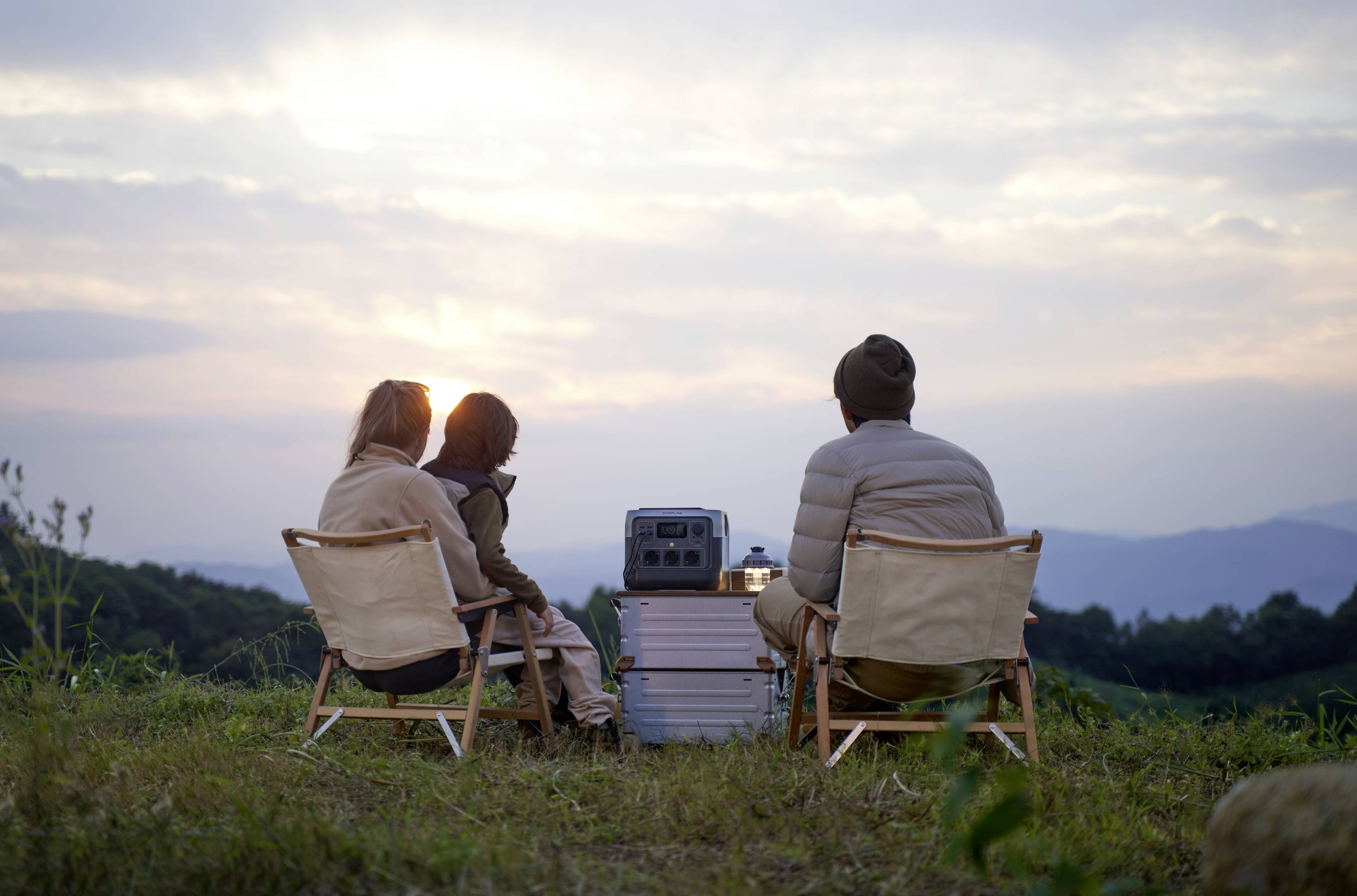 Drei Personen sitzen auf Campingstühlen und blicken bei Sonnenuntergang über eine Landschaft. Ein tragbarer Energiegenerator steht daneben.