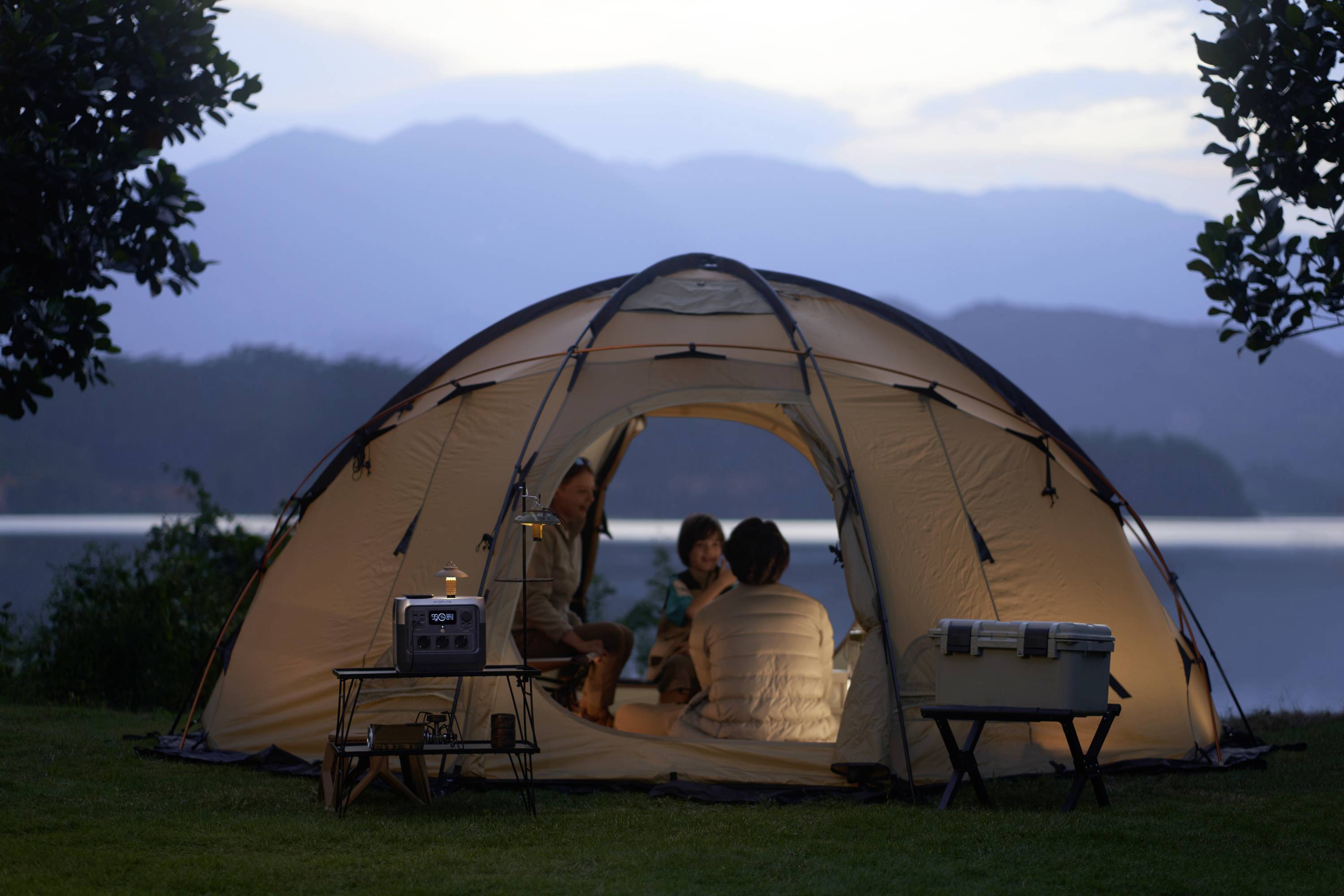 Drei Personen sitzen in einem beleuchteten Zelt, umgeben von Bäumen, mit Blick auf einen See und Berge im Hintergrund bei Abenddämmerung.