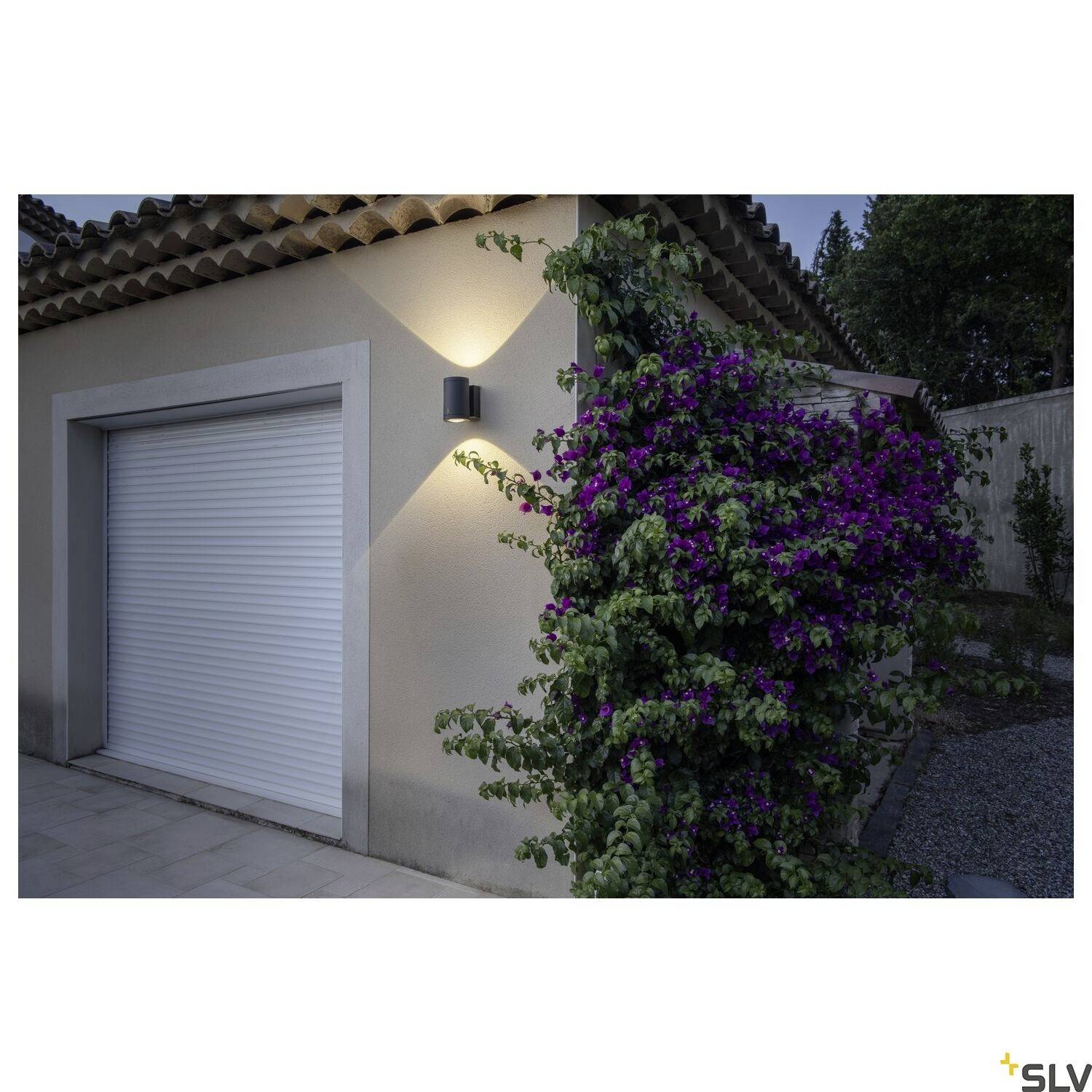 Außenwand mit Beleuchtung neben einer Garage. Efeu-Ranke mit lila Blüten wächst an der Wand empor.