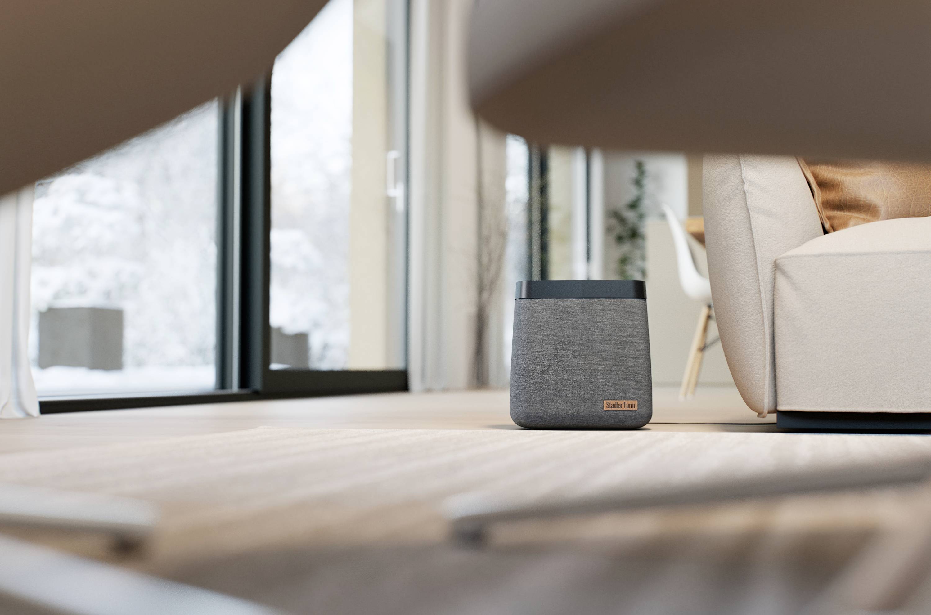 Ein grauer Lautsprecher steht auf Holzboden in einem modernen Wohnzimmer, mit großen Fenstern, die auf eine verschneite Landschaft blicken.