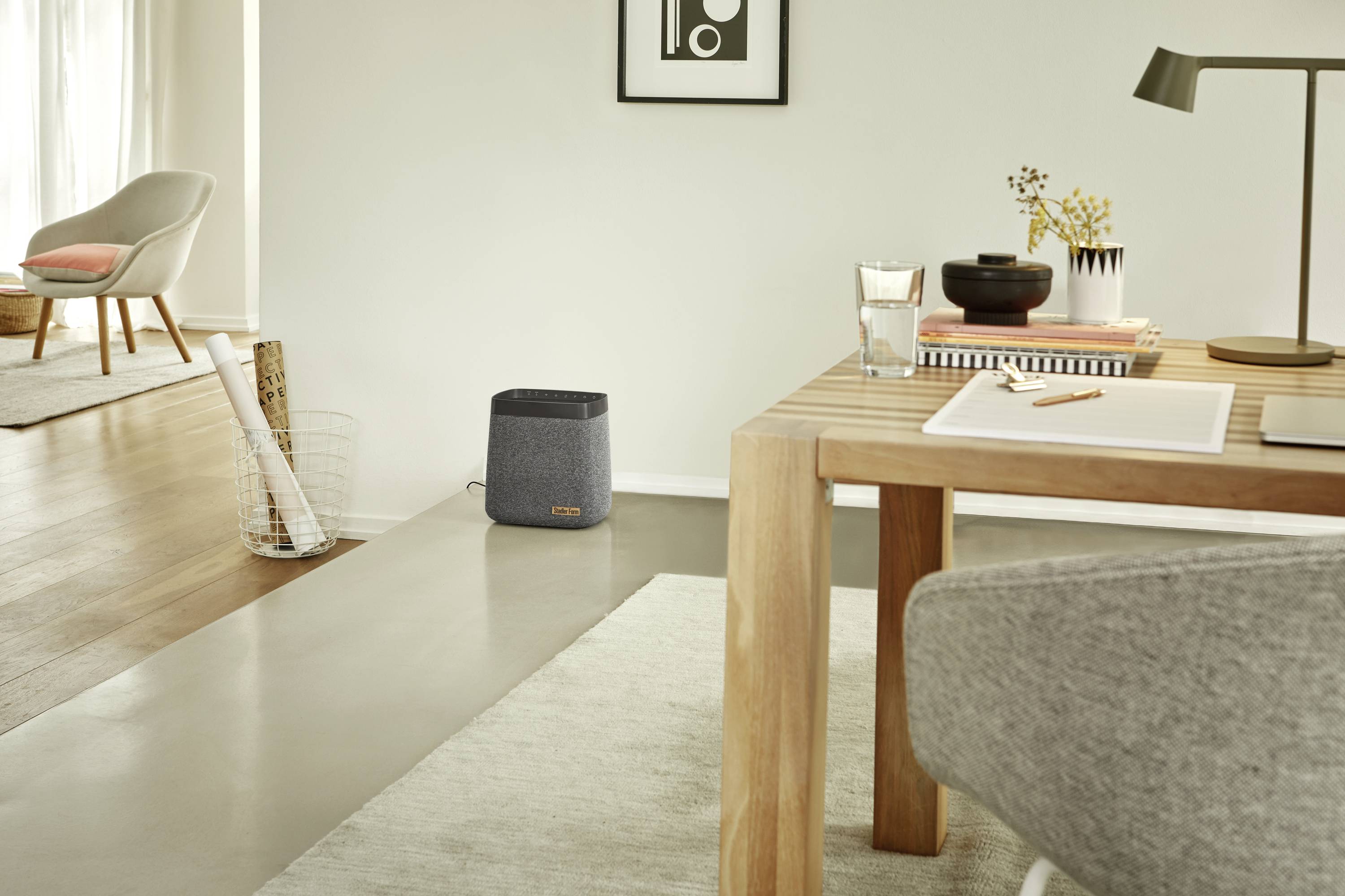 Ein modernes Wohnzimmer mit einem Tisch, auf dem Bücher und eine Schreibtischlampe stehen. Im Hintergrund ein Sessel und ein Lautsprecher.