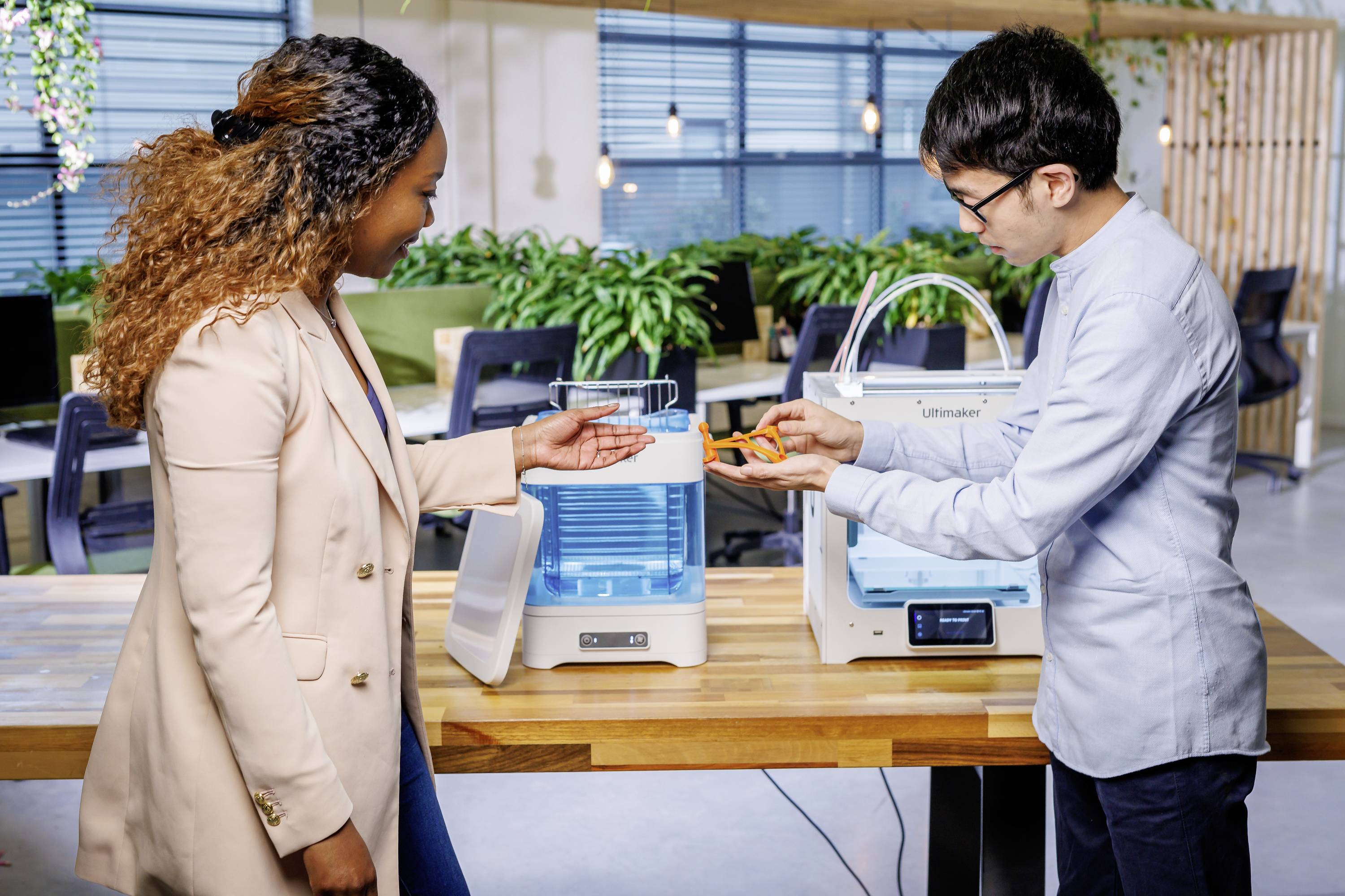 Eine Frau und ein Mann diskutieren in einem modernen Büro über 3D-gedruckte Objekte. Im Hintergrund sind 3D-Drucker zu sehen.